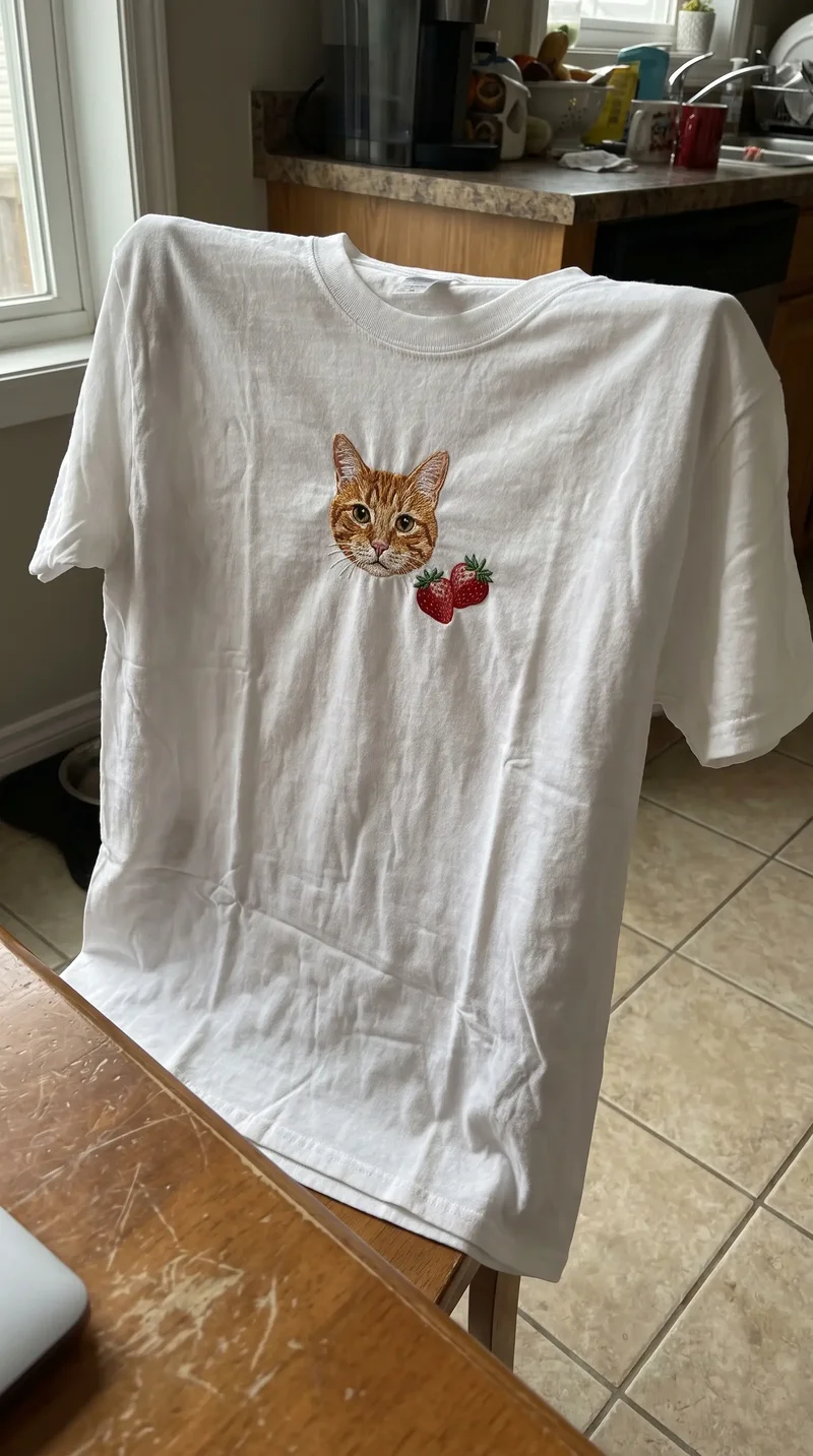 Tricou alb cu o pisică portocalie brodată și două căpșuni, pe spătarul unui scaun