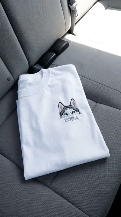Tricou alb împăturit pe bancheta mașinii, cu un husky brodat și numele ZORA dedesubt