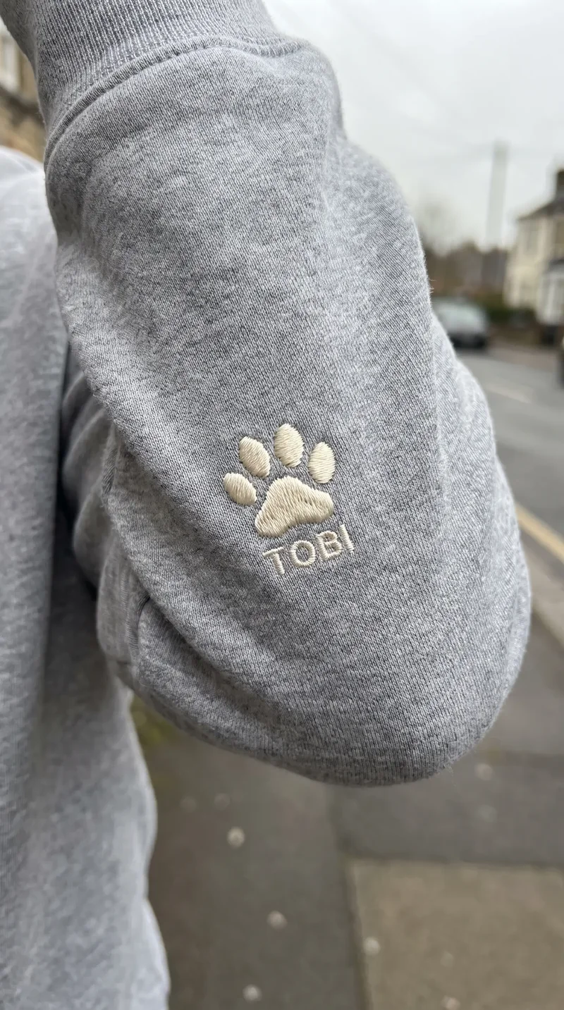 Lăbuță brodată și numele TOBI pe mâneca unui hanorac gri